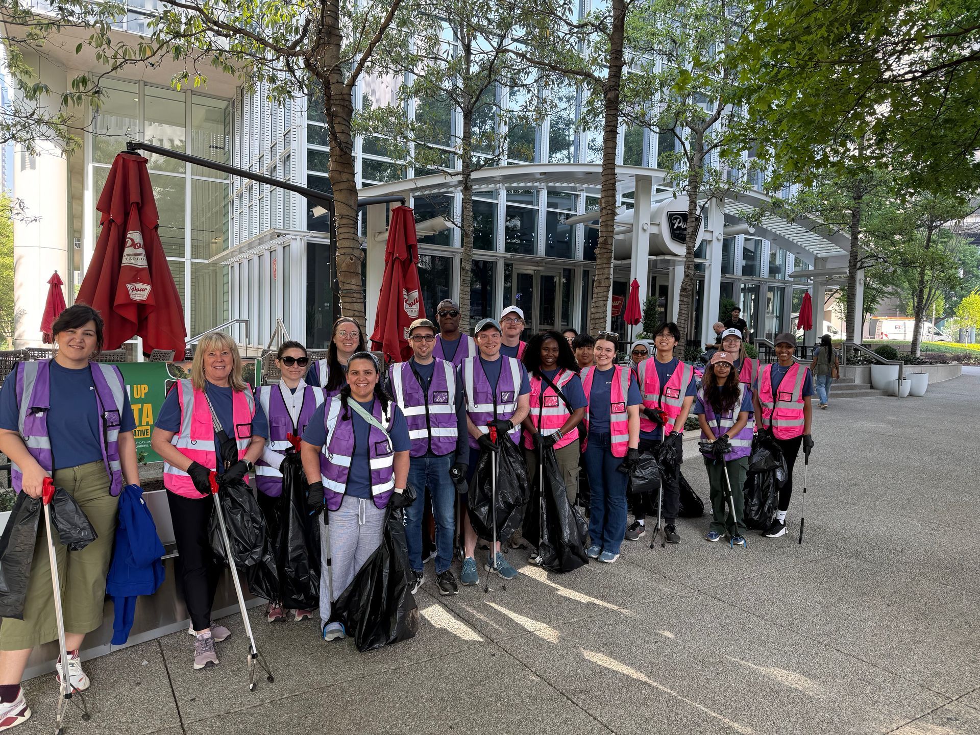 Group of volunteers in pink safety vests and purple shirts standing outdoors with trash bags and litter grabbers