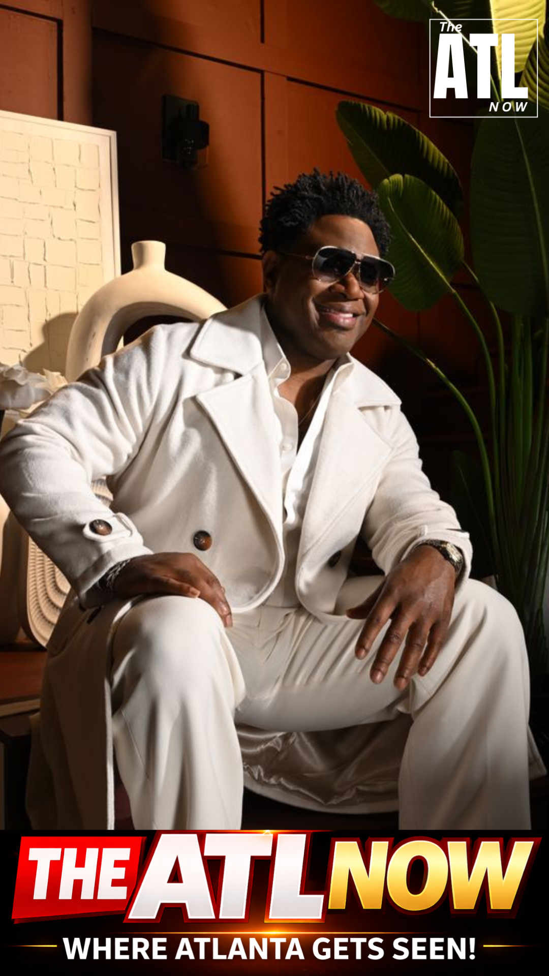 Man in white suit and sunglasses seated indoors beside a lamp and plant, with “THE ATL NOW” text.