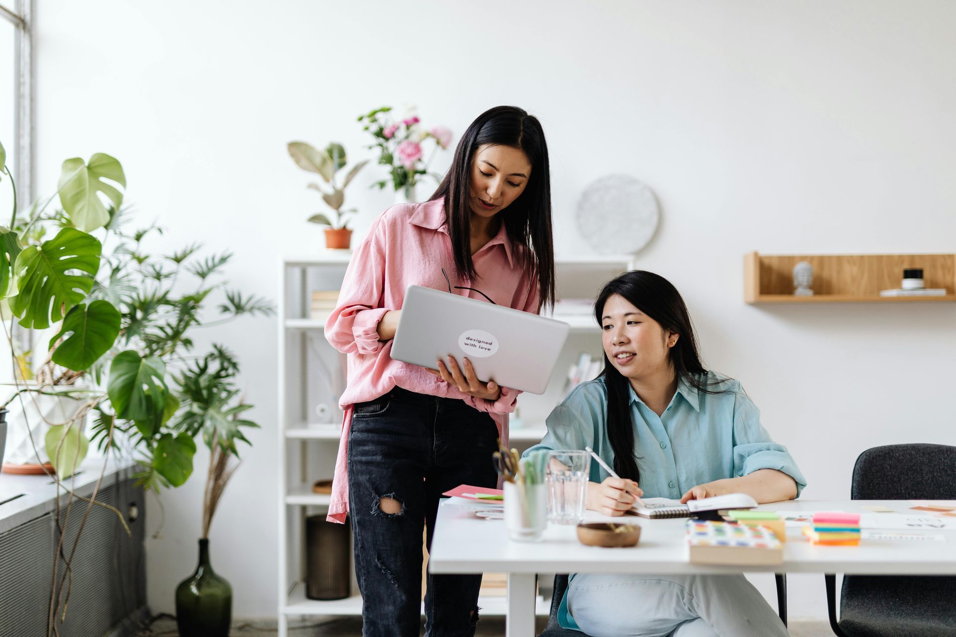 Two people in an office looking at a laptop. One is standing, holding the laptop; the other is seated, writing.
