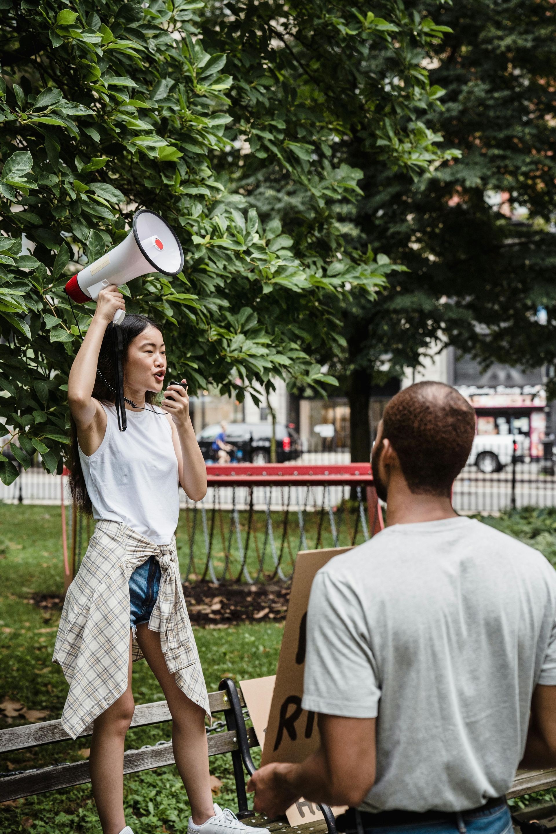 Woman with megaphone on bench protests with a sign held by a person nearby, in a park.