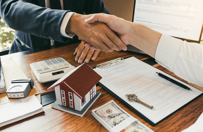 A man and woman are shaking hands over a table with a model house and money.