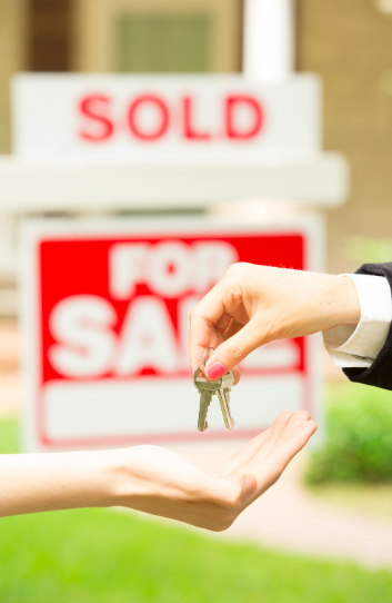 A person is handing keys to another person in front of a sold for sale sign
