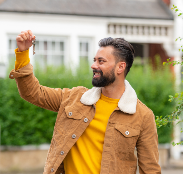 A man in a brown jacket is holding a key in front of a house.