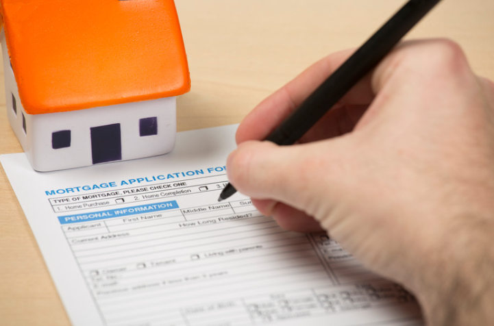 A person is writing on a mortgage application form with a model house in the background.