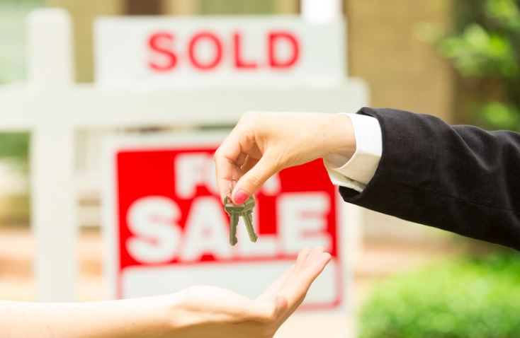 A person is handing a key to another person in front of a sold sign.