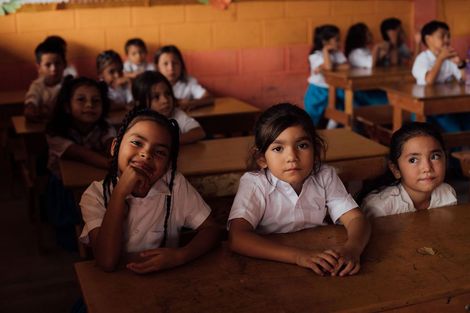 Children in white shirts and dark skirts stand in a line outdoors, smiling in bright sunlight.
