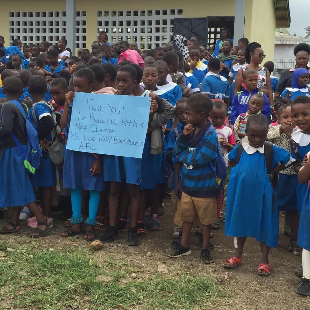Group of schoolchildren in blue uniforms gathered outdoors, holding a handwritten sign in front of a building.