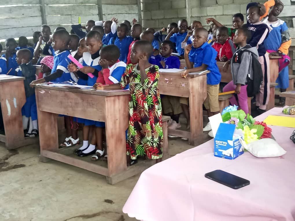 Children seated at wooden desks in a classroom, with one adult standing nearby and papers on a table in front.