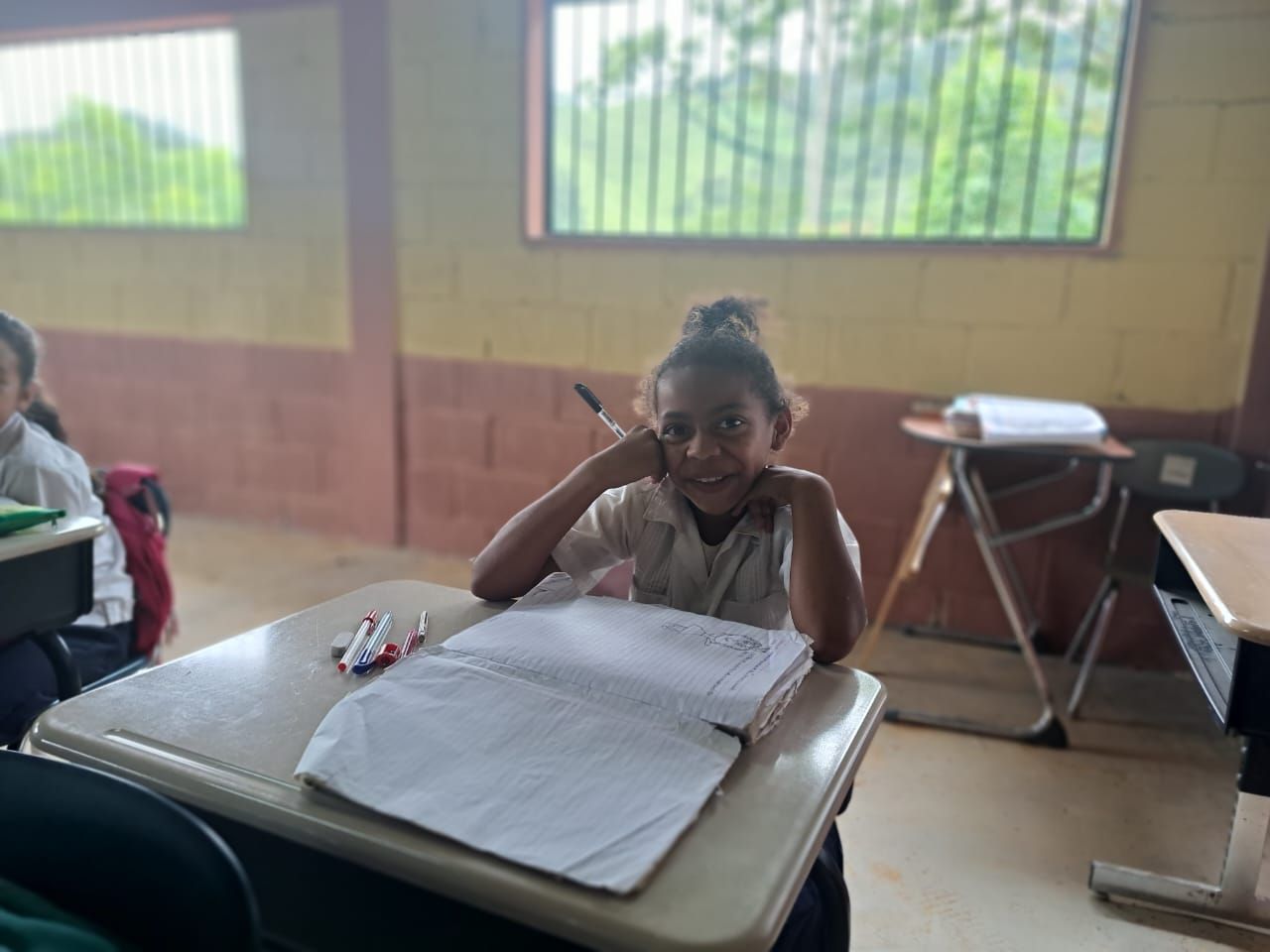 Student seated at a desk in a classroom, smiling with a notebook open in front of her.