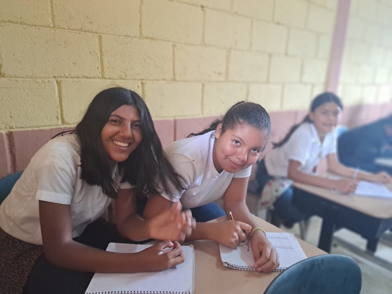 Two students smiling and writing at desks in a classroom