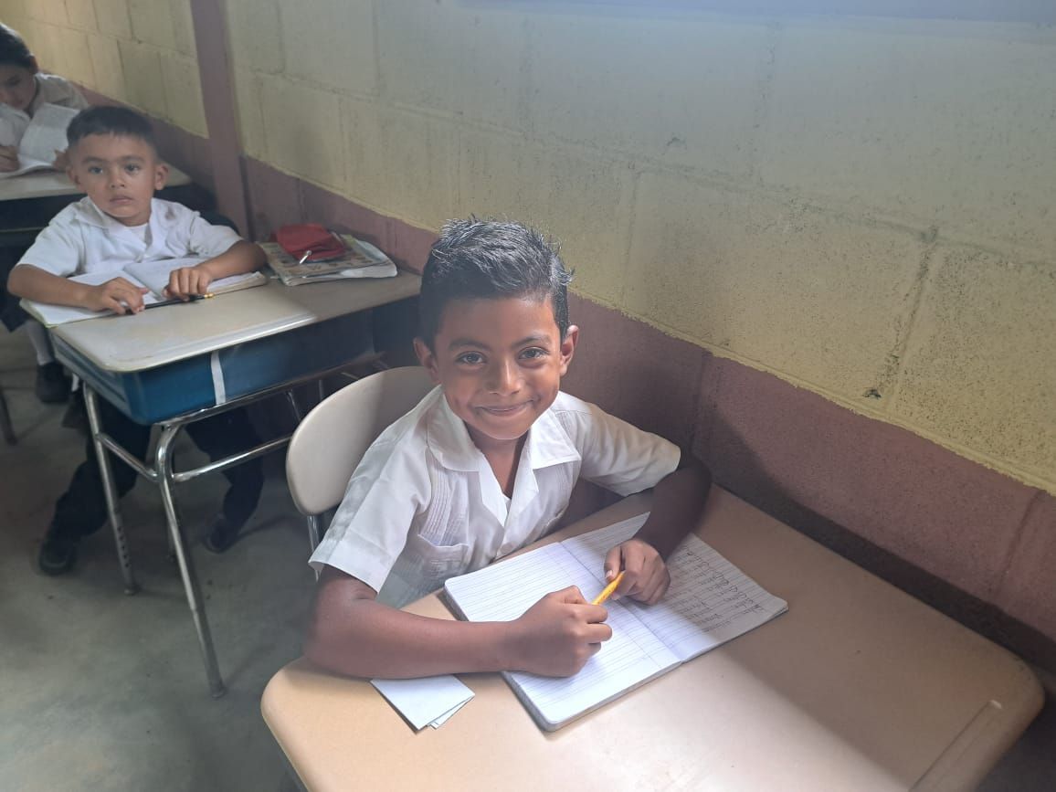 Two students in white uniforms sit at desks in a classroom, writing in notebooks.