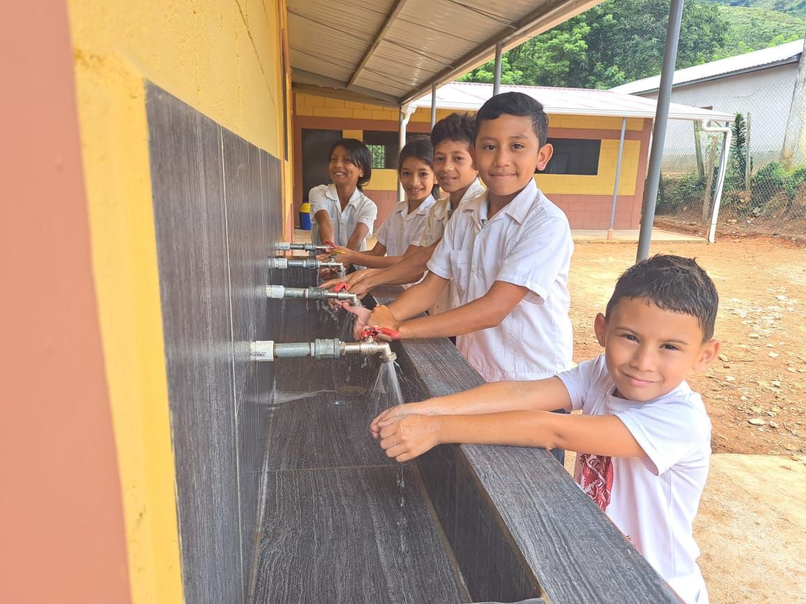 Children washing hands at a row of outdoor sinks under a covered walkway.