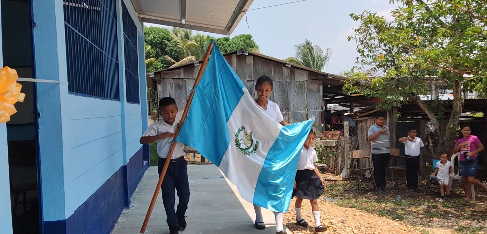 People carrying a blue-and-white flag outside a blue building in a rural village