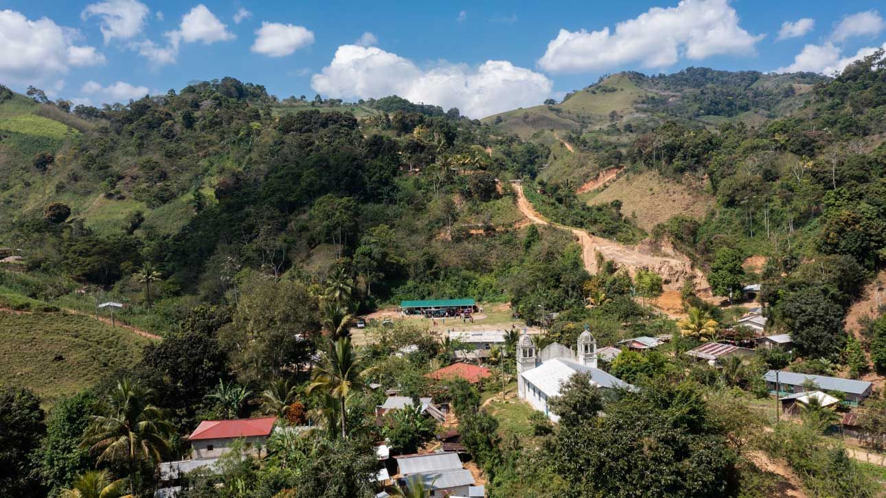 Rural hillside village with scattered houses, green trees, and a dirt road winding up the mountain under a blue sky.