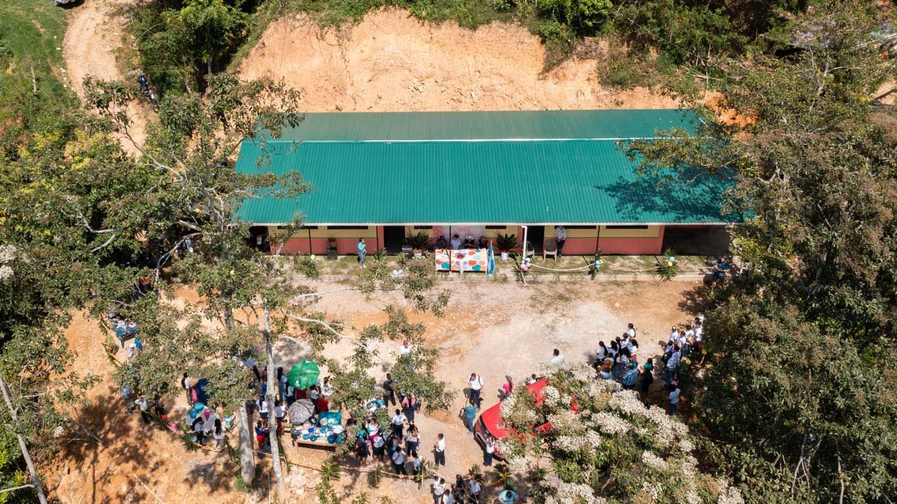 Aerial view of a long building with a teal roof, surrounded by a crowd and trees in a clearing.