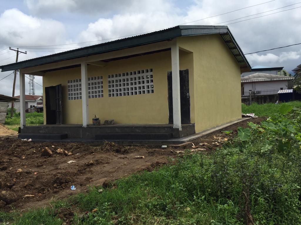 Small yellow house with dark roof beside a muddy yard and green vegetation under cloudy skies