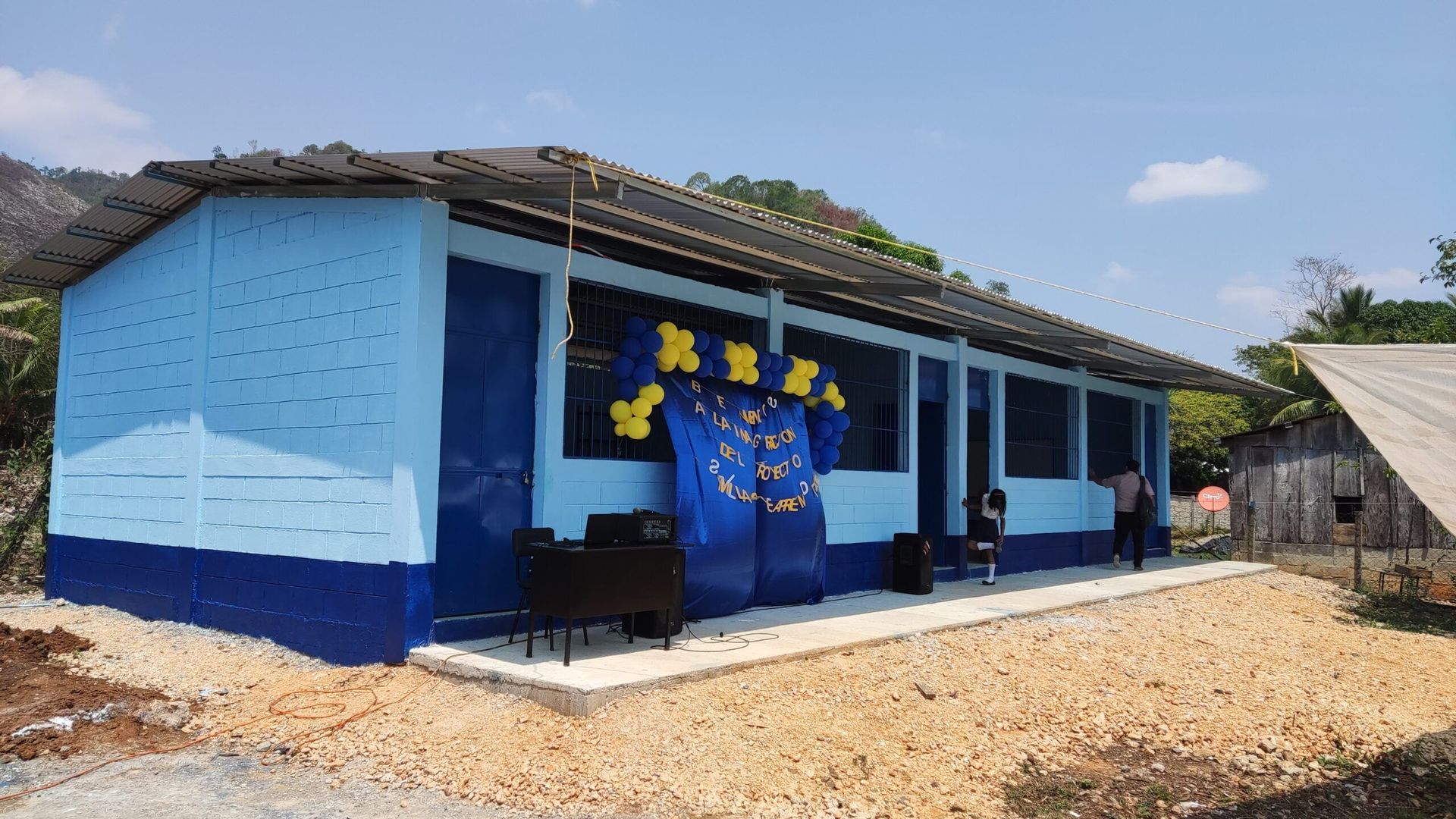 Blue rural classroom building with a covered porch and star decorations, on a dirt lot under a sunny sky