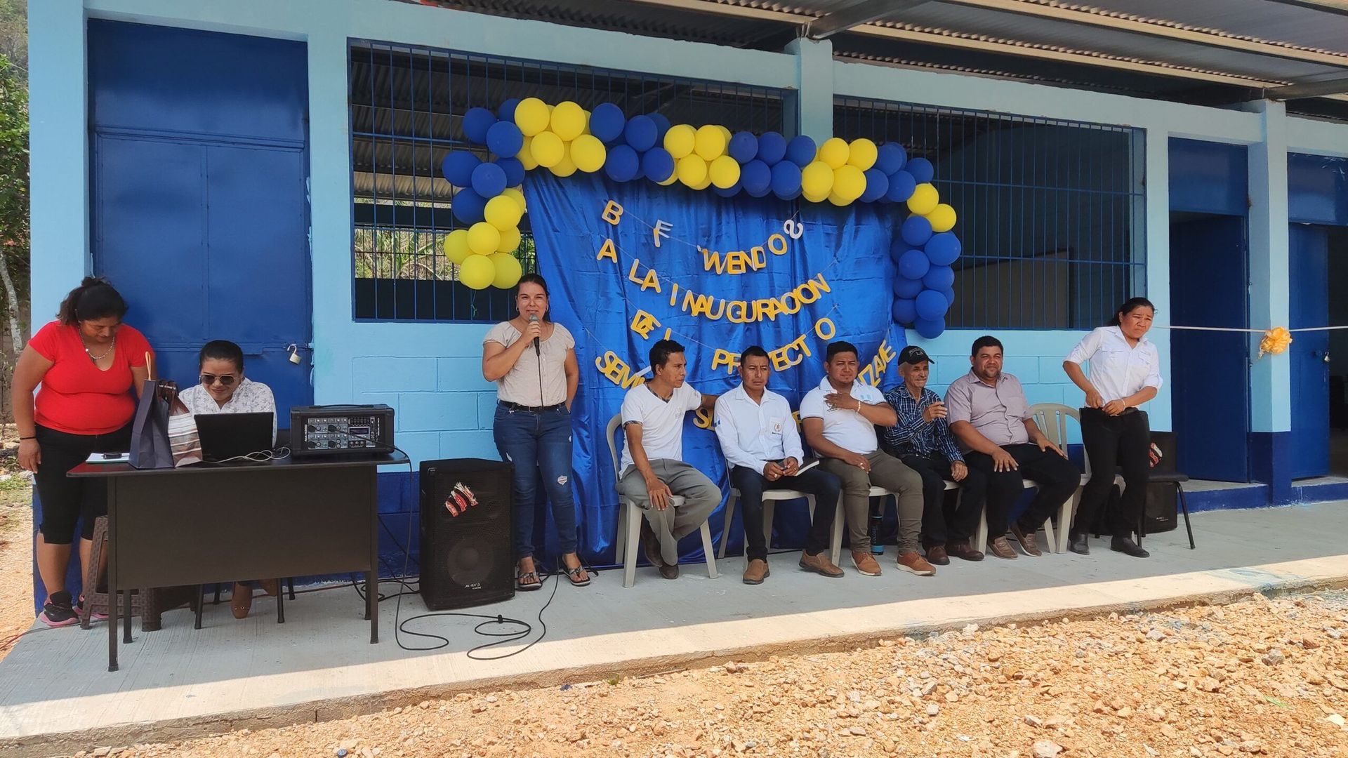 Group seated on stage with blue and yellow balloon backdrop at an outdoor ceremony