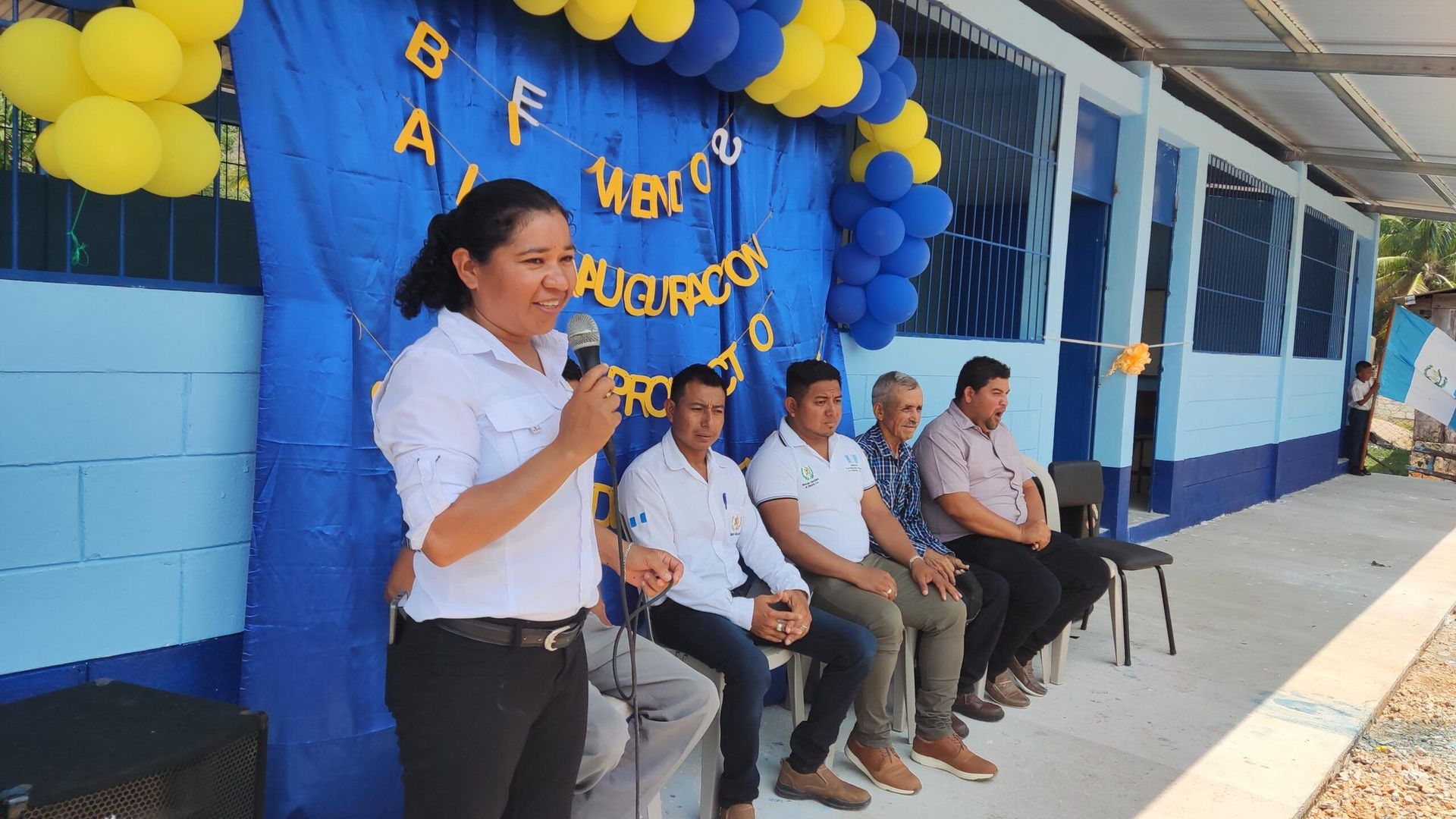 Woman speaking at a microphone beside seated people under blue and yellow balloons outside a building