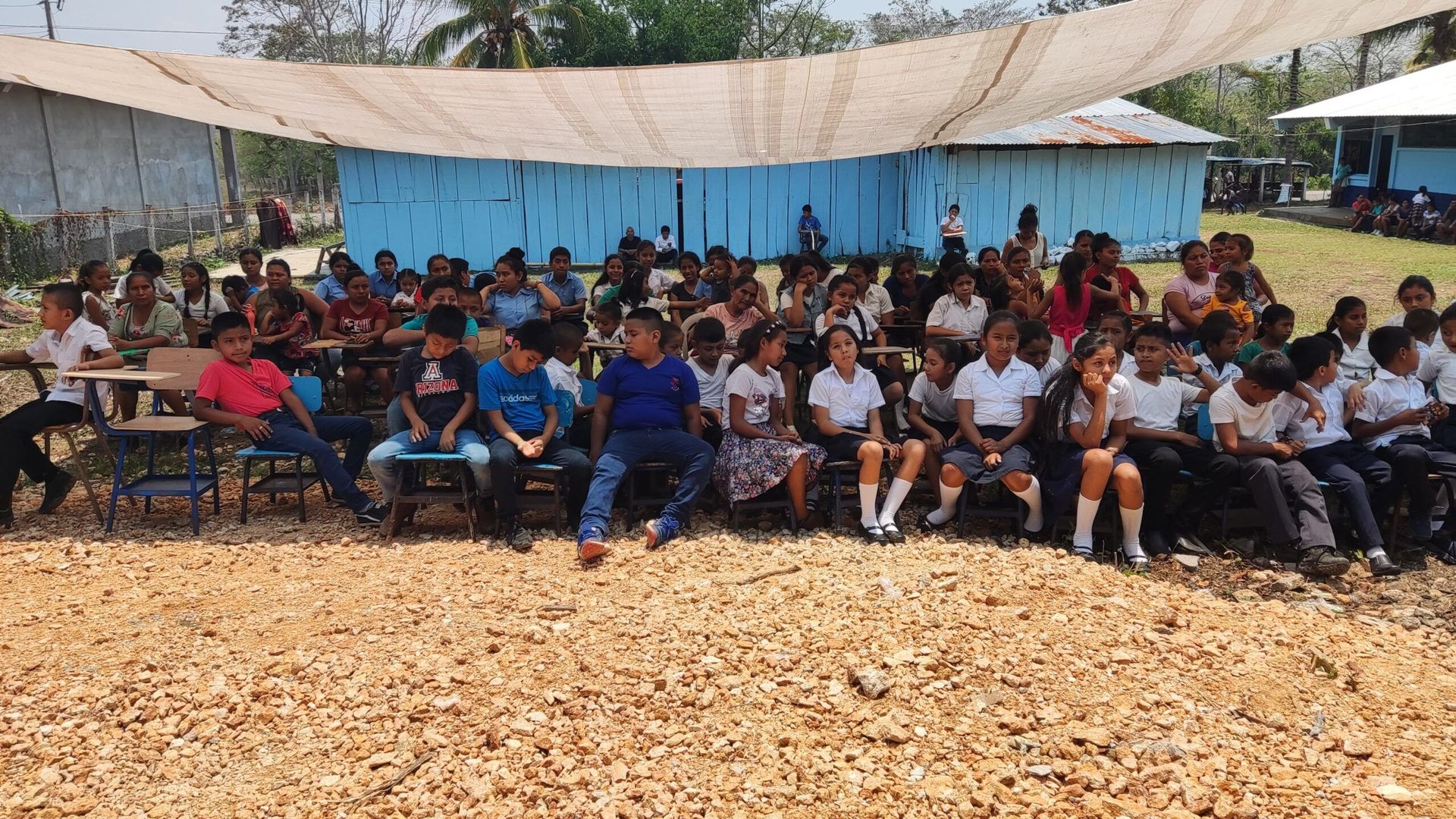 Large group seated under a tent at an outdoor event, with blue backdrop and gravel ground.