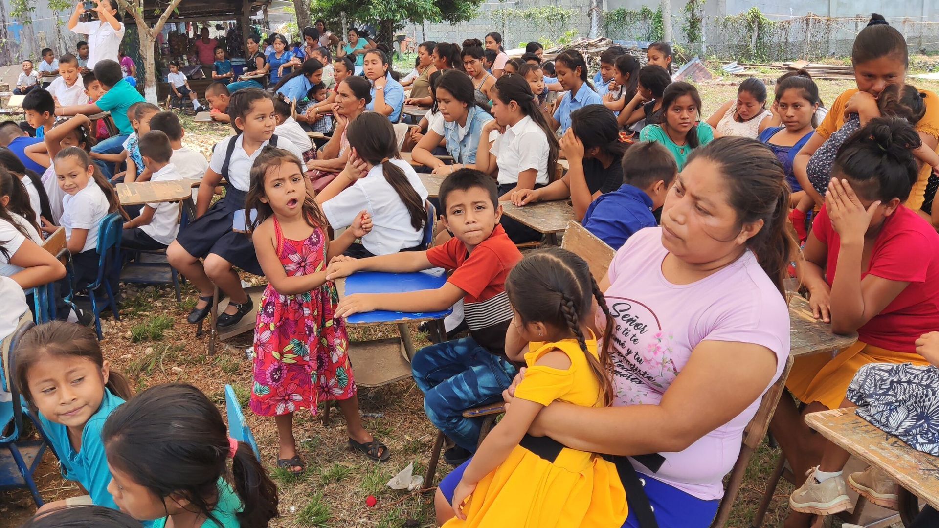 Crowded outdoor gathering with children and adults sitting together, many holding plates and food.
