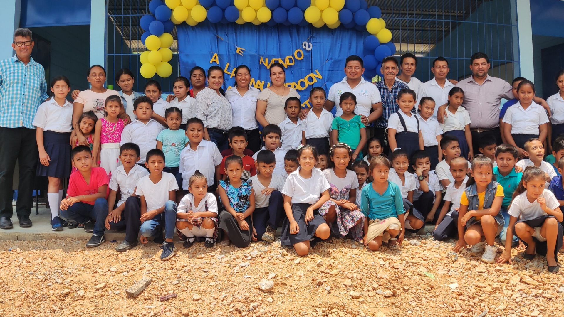 Group of children and adults posing outdoors in front of a blue-and-yellow balloon-decorated school entrance