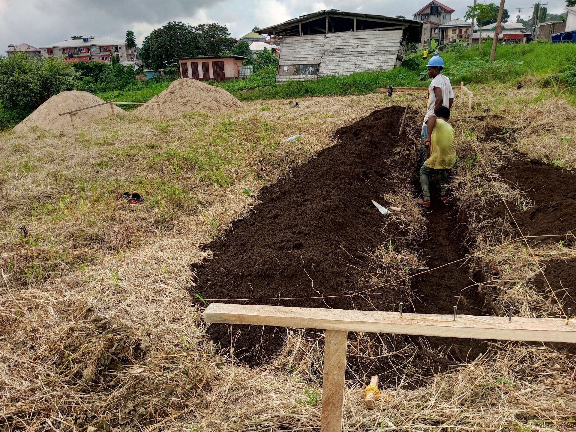 Workers digging a long trench in a grassy field with piles of soil and a wooden barrier in the foreground