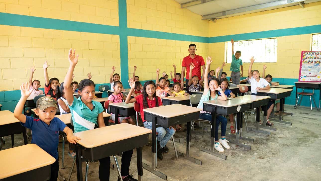 Classroom of children seated at desks with hands raised, with teachers standing at the front.