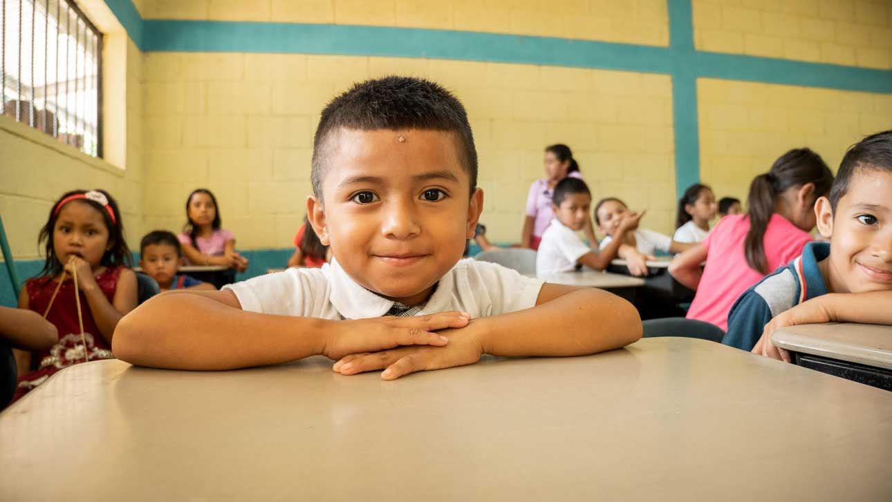 Child smiling at a classroom desk, leaning forward with hands folded, with other students in the background