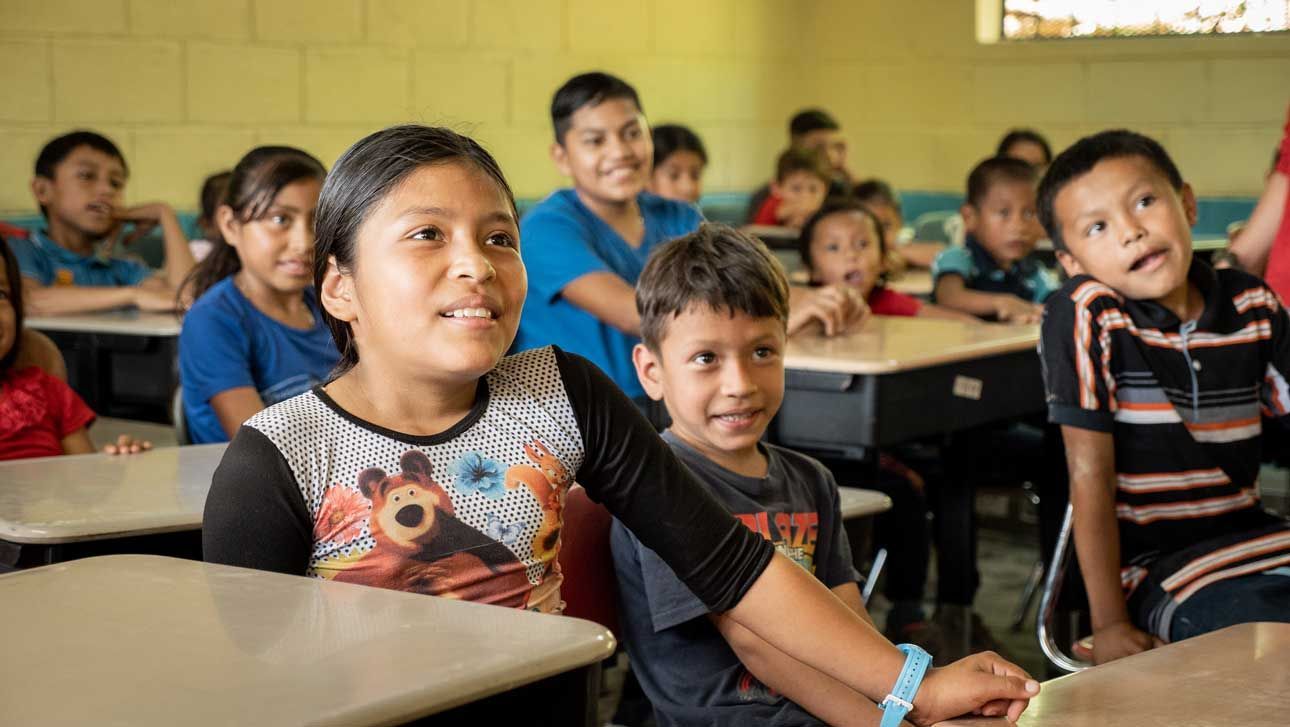 Children seated at desks in a classroom, smiling and looking toward the front.