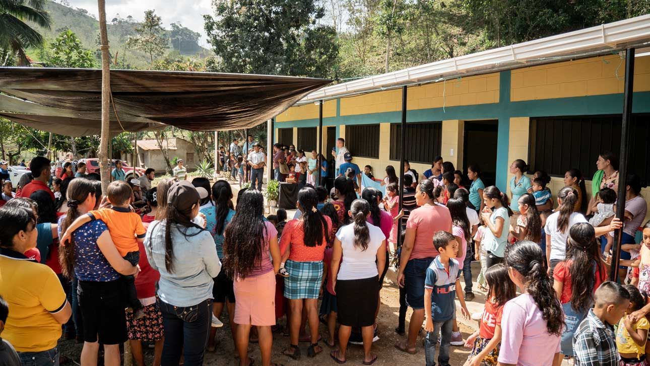 Crowd gathered outside a school building under a canopy, with people standing closely together in a courtyard.