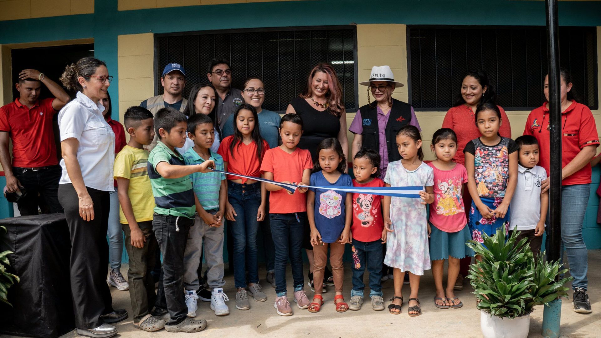 Group of people posing outside a building, with children in front holding a red ribbon.