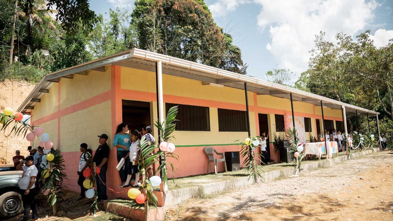 Open-air community building with peach walls, people gathered outside, and potted plants along the porch
