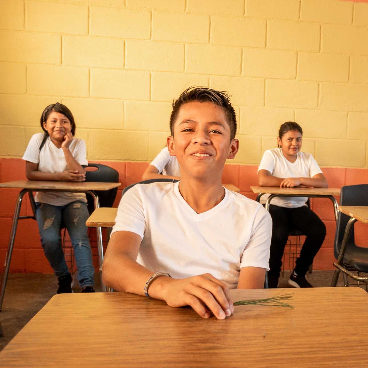 Three students sit at desks in a classroom with yellow walls, facing the camera.