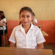 Student seated at a classroom desk, wearing a white shirt, looking at the camera.