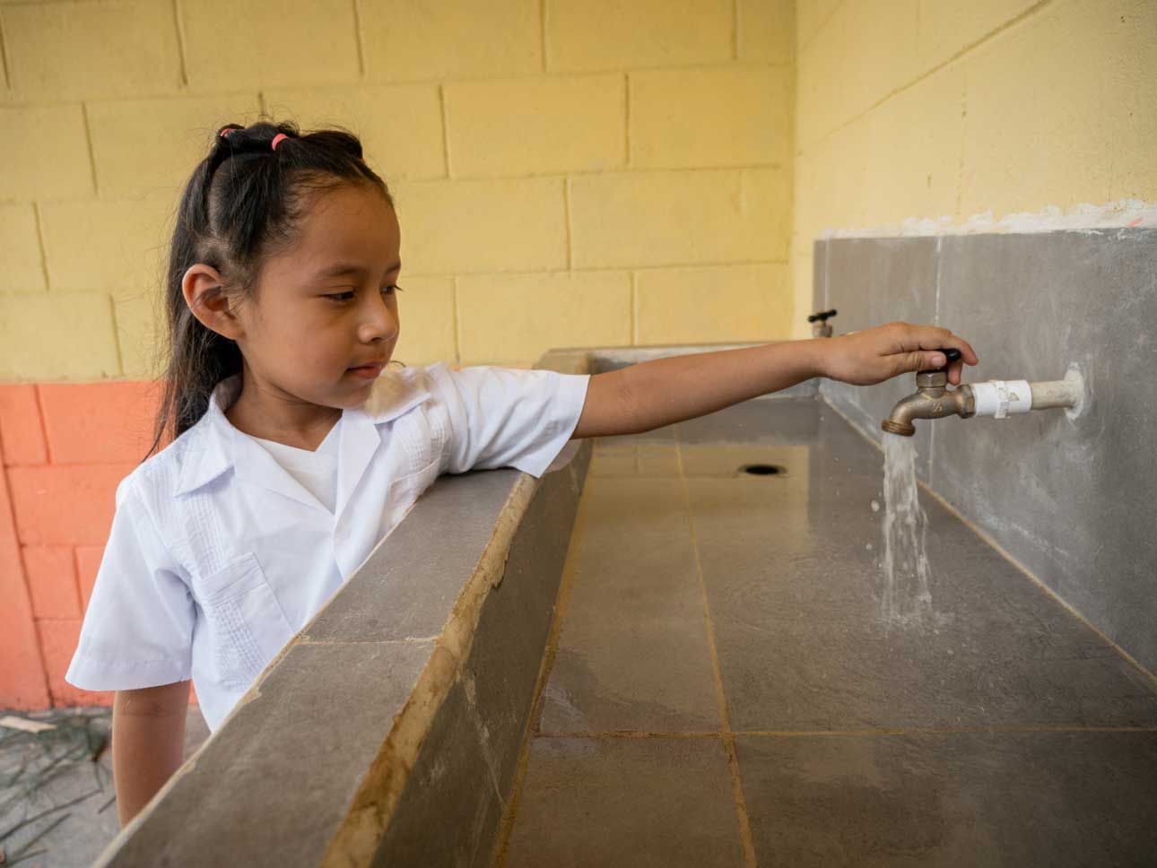 Child in white shirt turns on a faucet at an outdoor sink, water running.