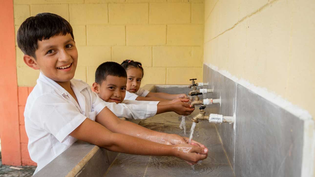 Children washing hands at a row of outdoor faucets, smiling in a school-style sink area.
