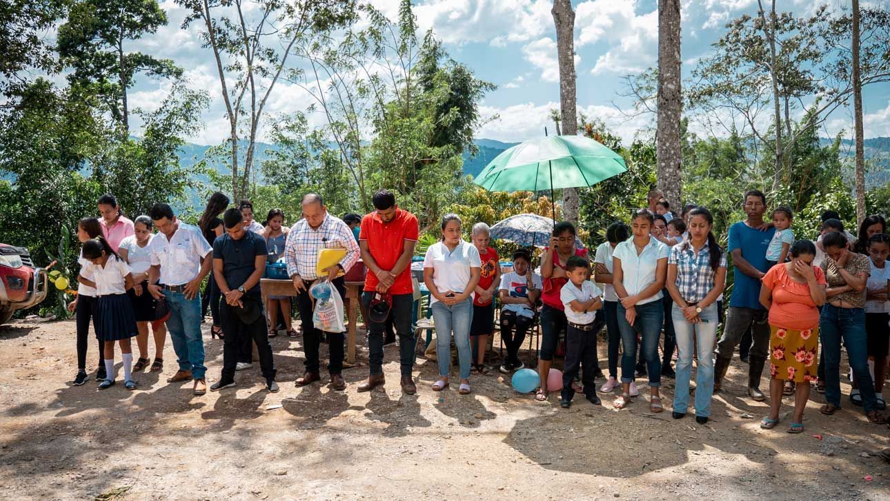 Group of people standing outdoors in a line, holding tools and a green umbrella under trees