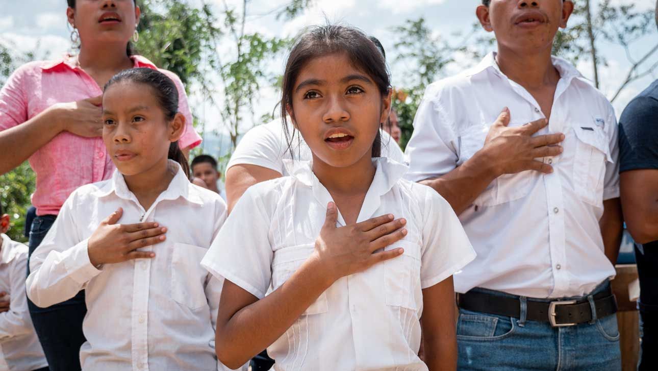 Students in white shirts stand with hands over hearts outdoors during a ceremony