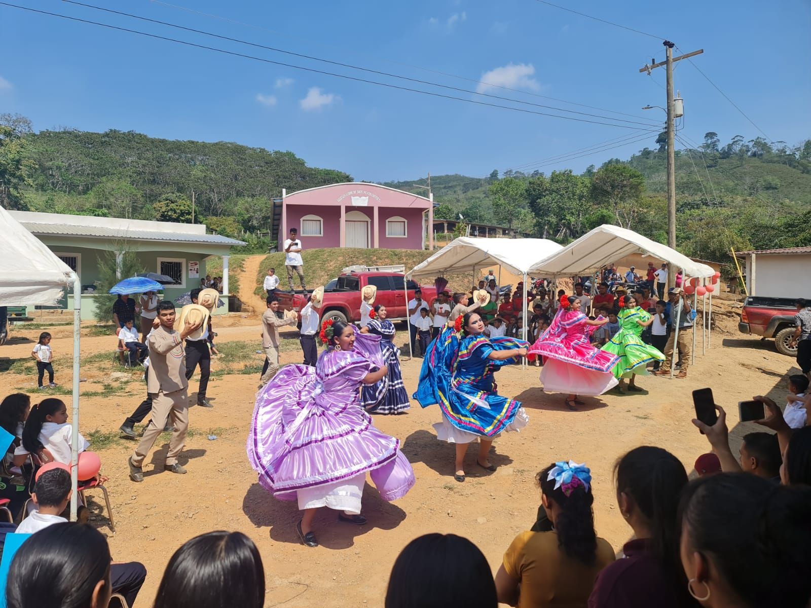 Colorful dancers perform outdoors at a community festival on a sunny dirt field.