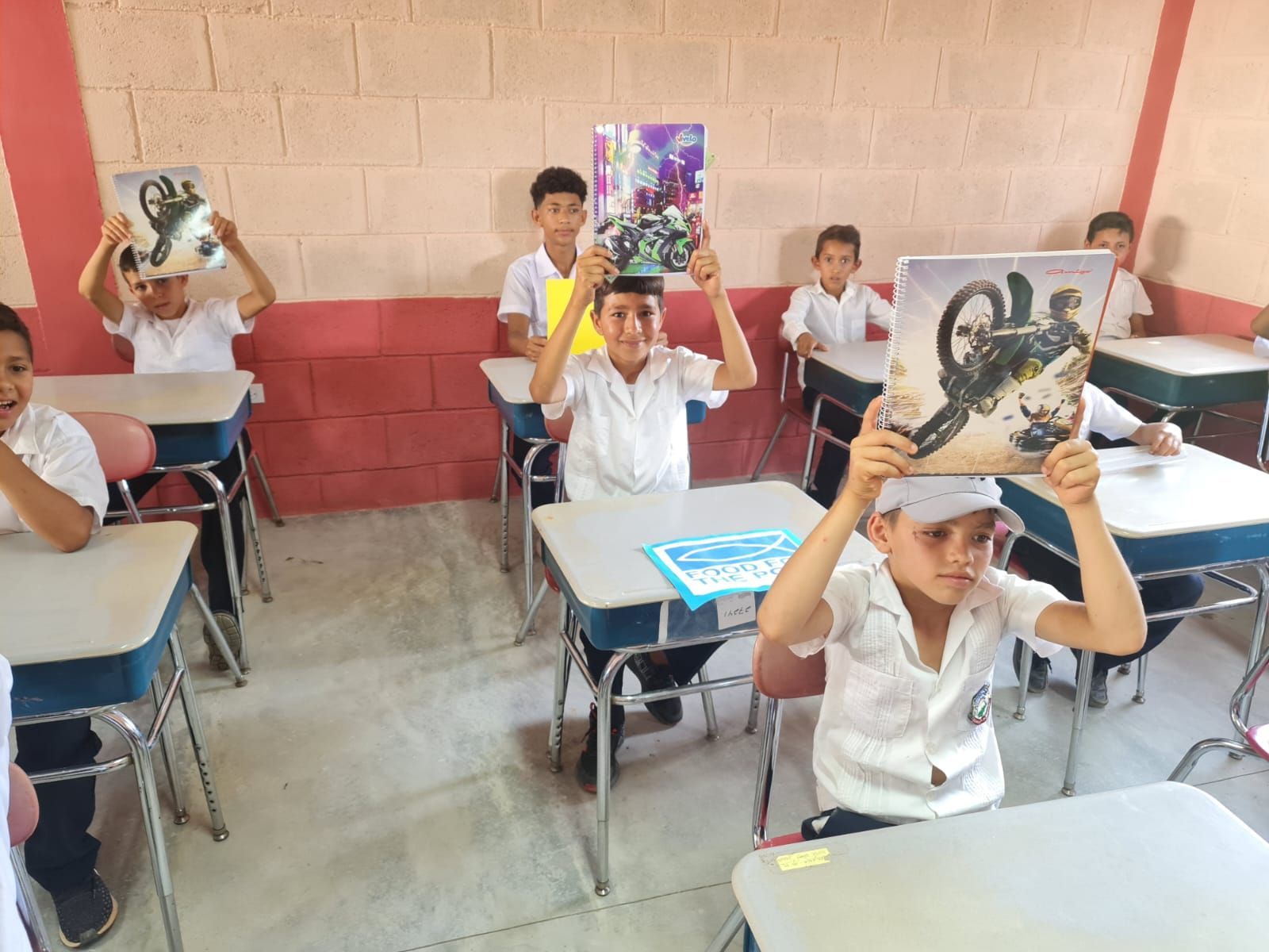 Students in a classroom hold up illustrated books while seated at desks.