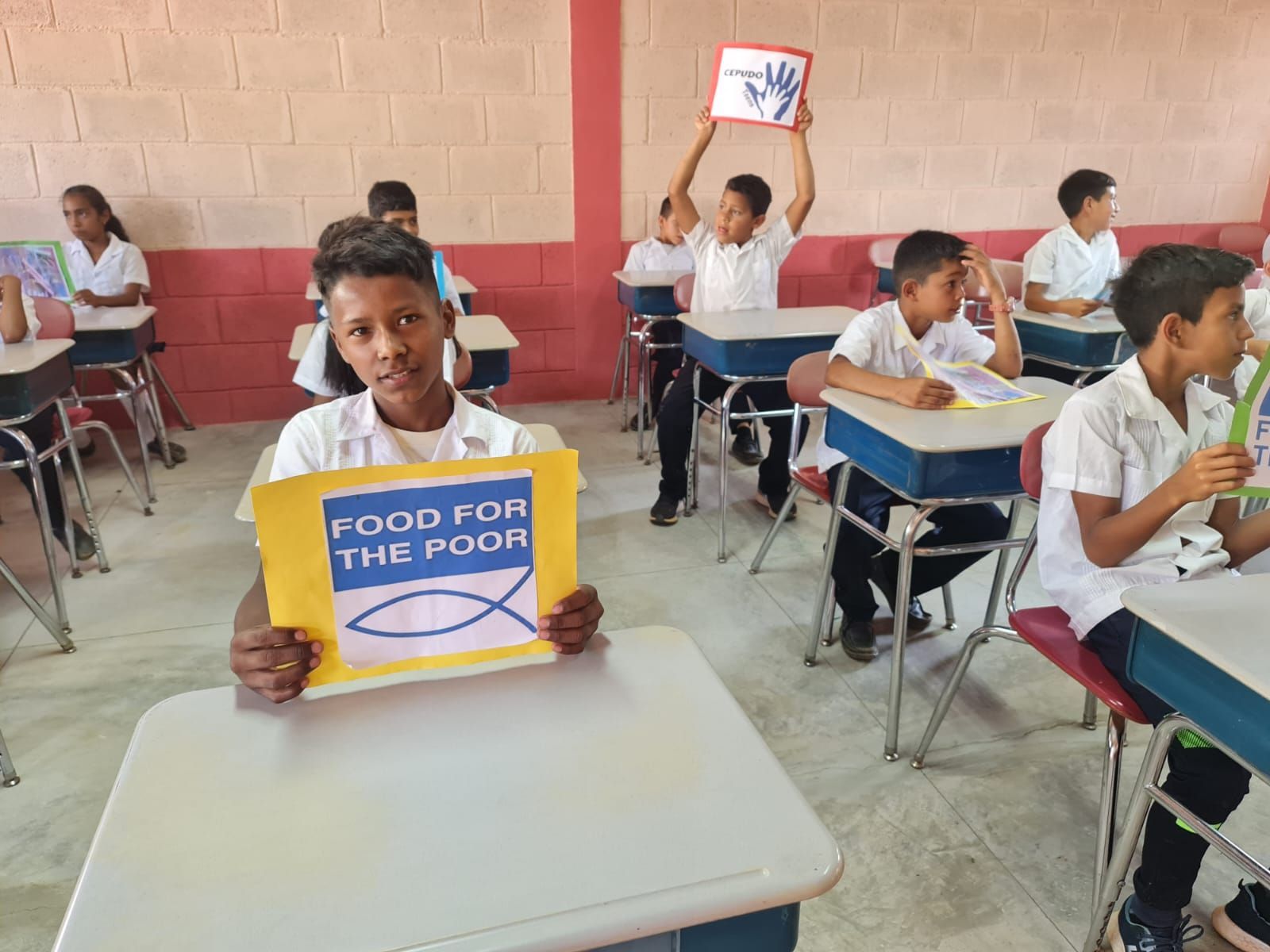 Student in classroom holding a blue sign that reads “FOOD FOR THE POOR.”