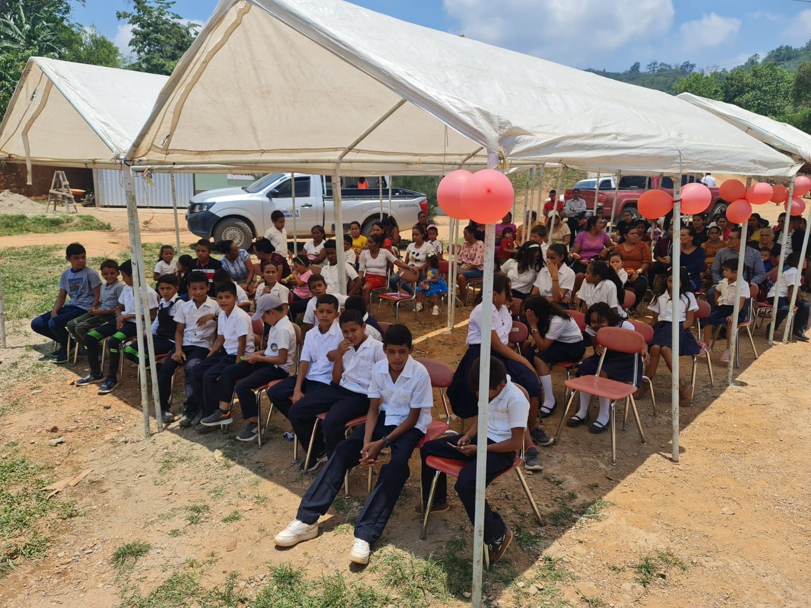 Students seated under a white tent at an outdoor gathering with red balloons and a dirt field nearby