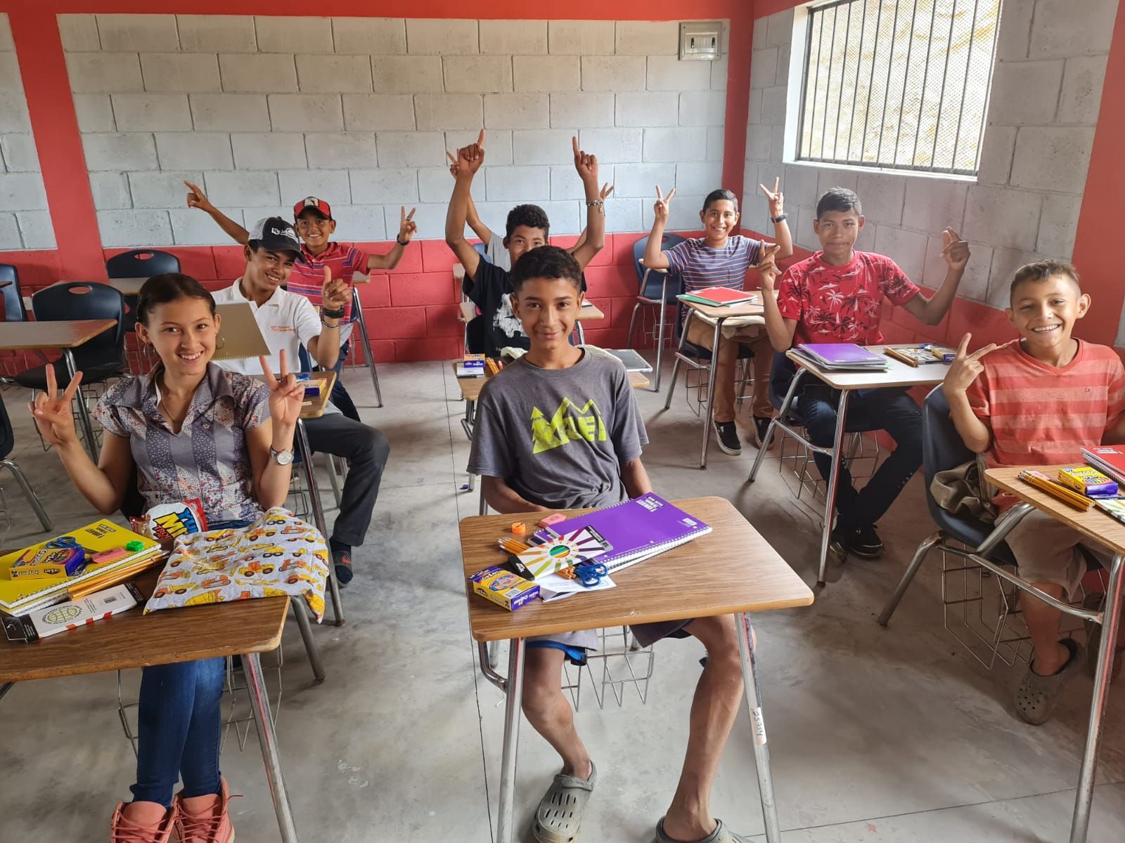 Students in a classroom with desks, colorful craft supplies, and several raising their hands.