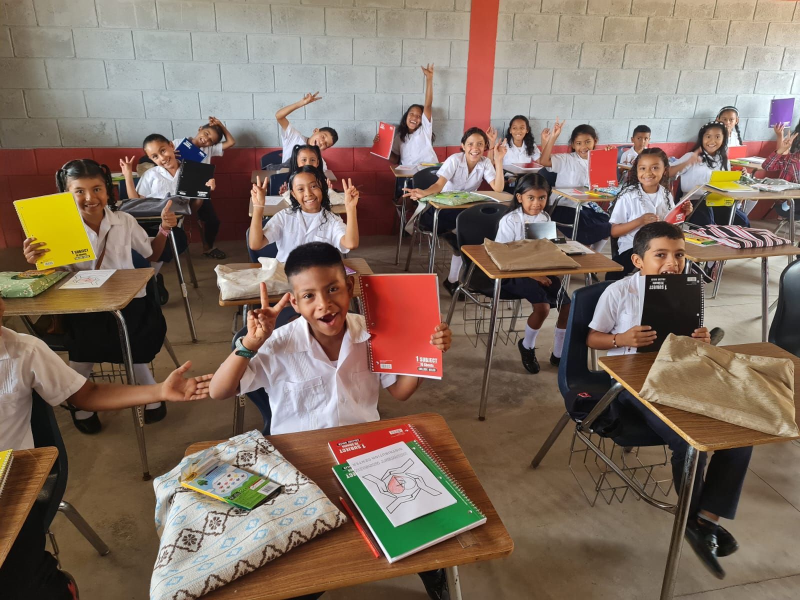 Students seated at desks in a classroom, holding up colorful notebooks and papers.