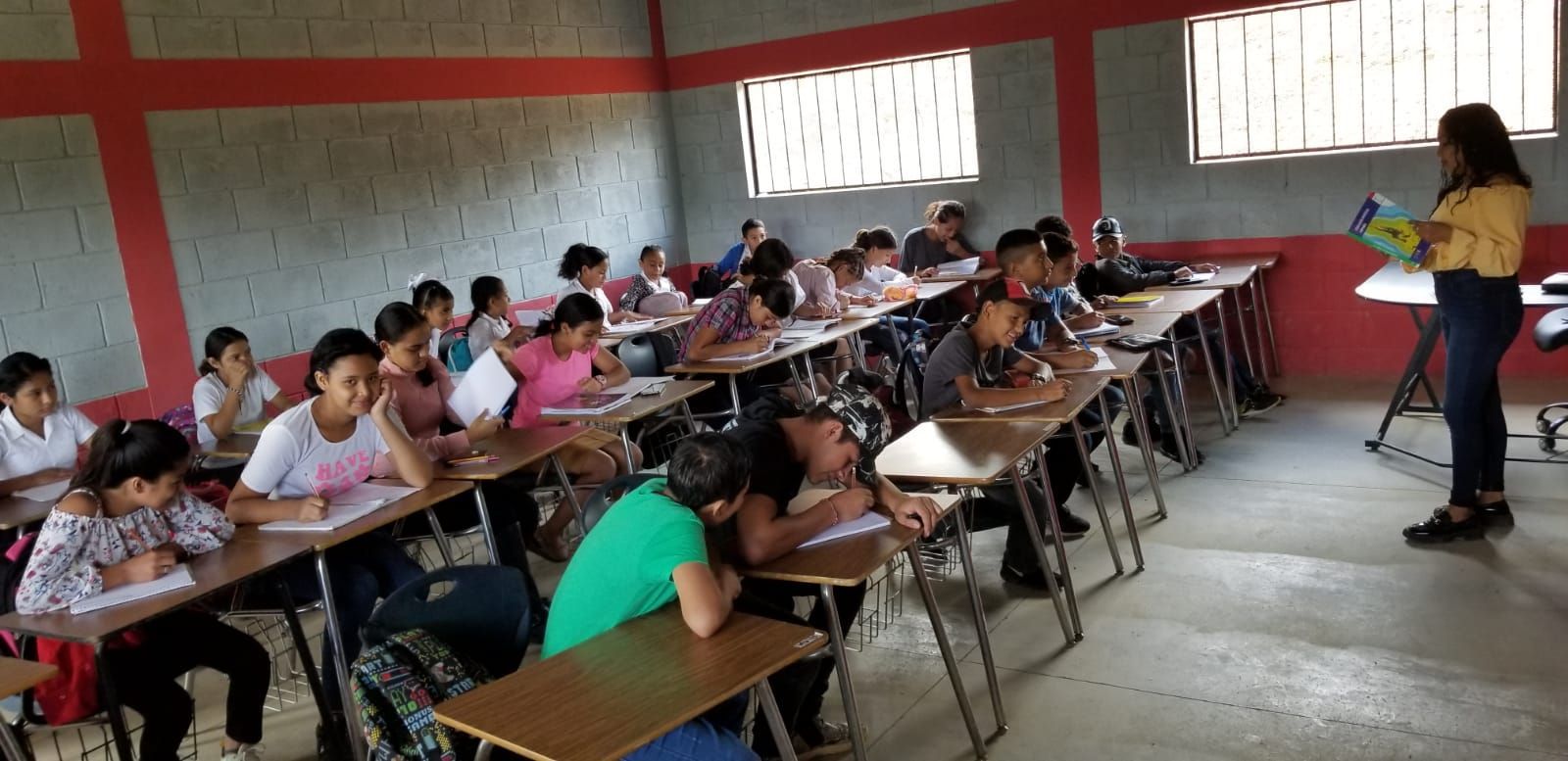 Students seated at desks in a classroom while a teacher stands at the front with a laptop