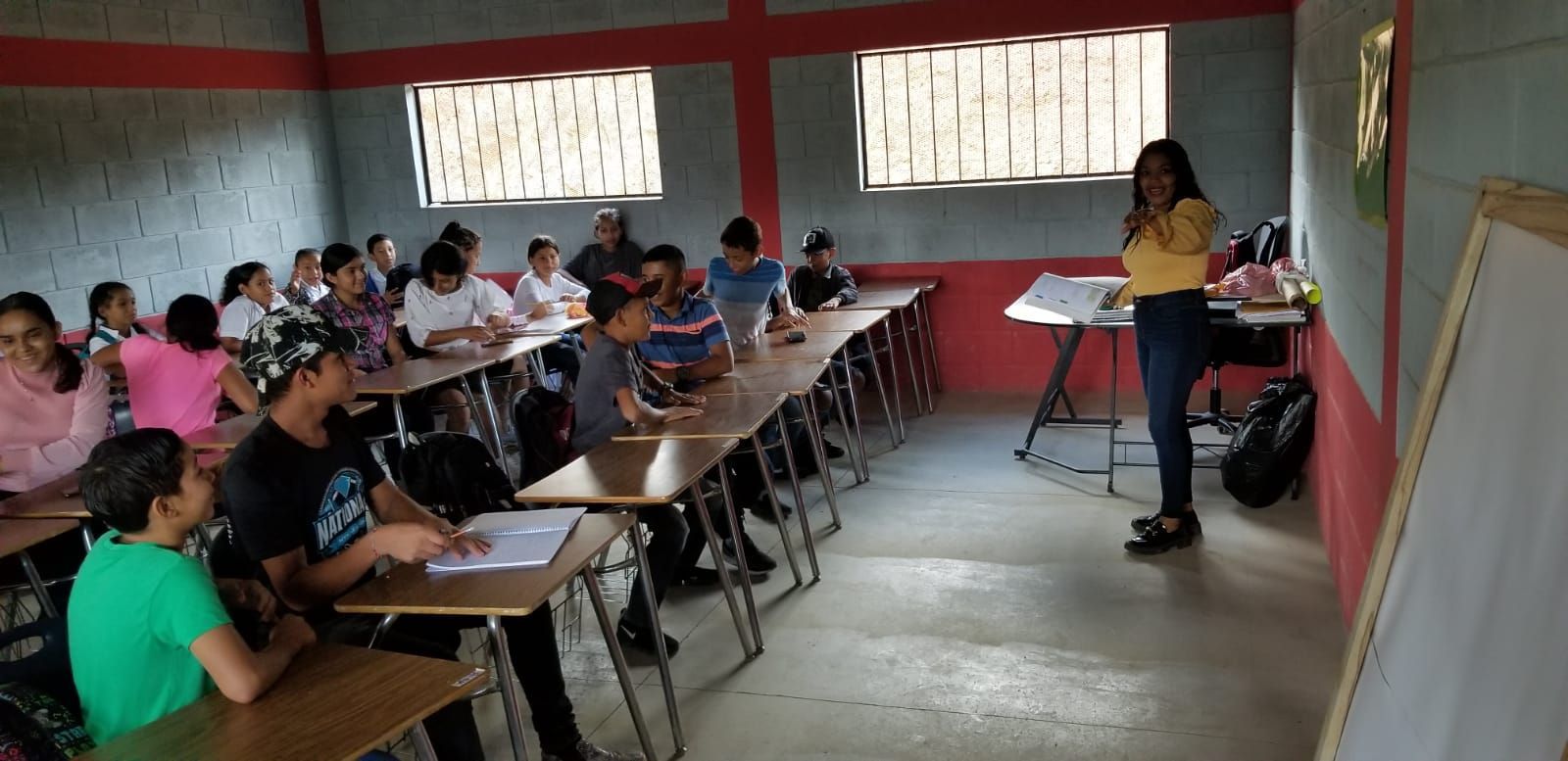 Classroom with students seated at desks, listening to a teacher near a whiteboard.