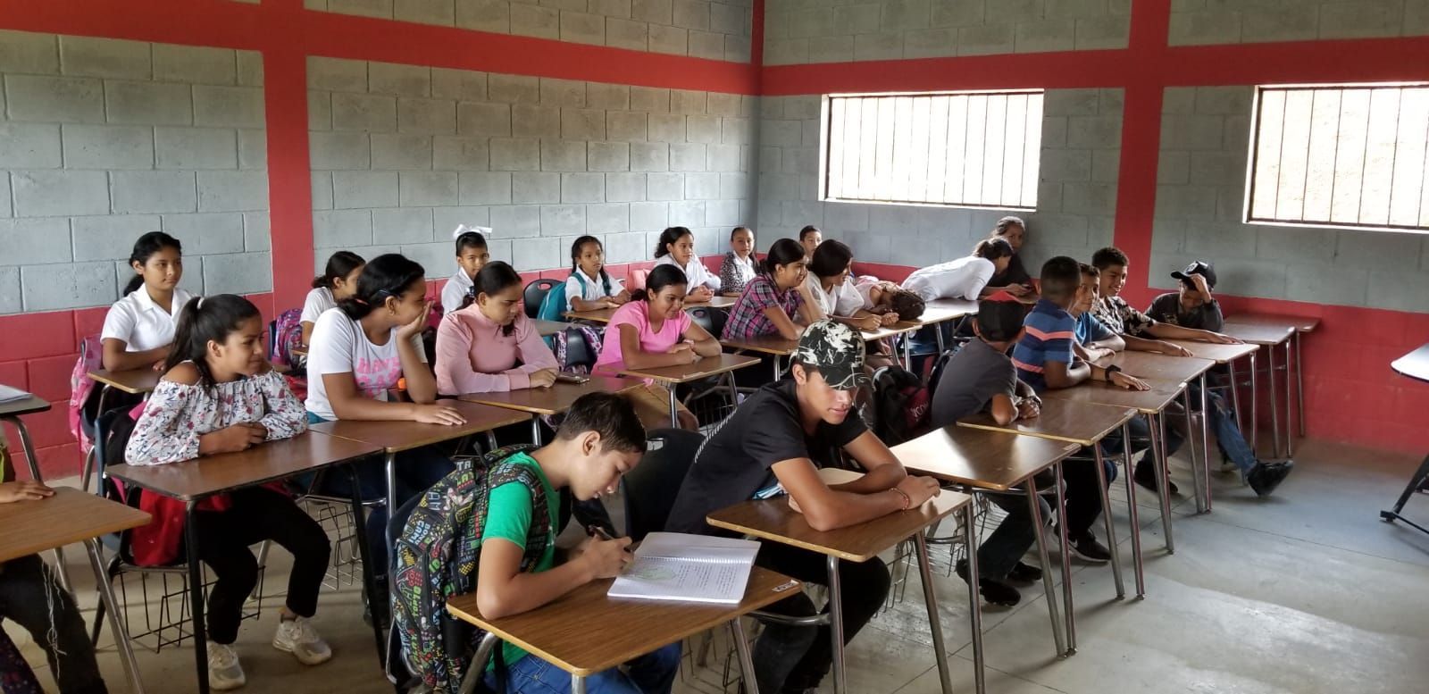 Students taking a written test in a classroom, seated at long desks with red walls and windows behind them
