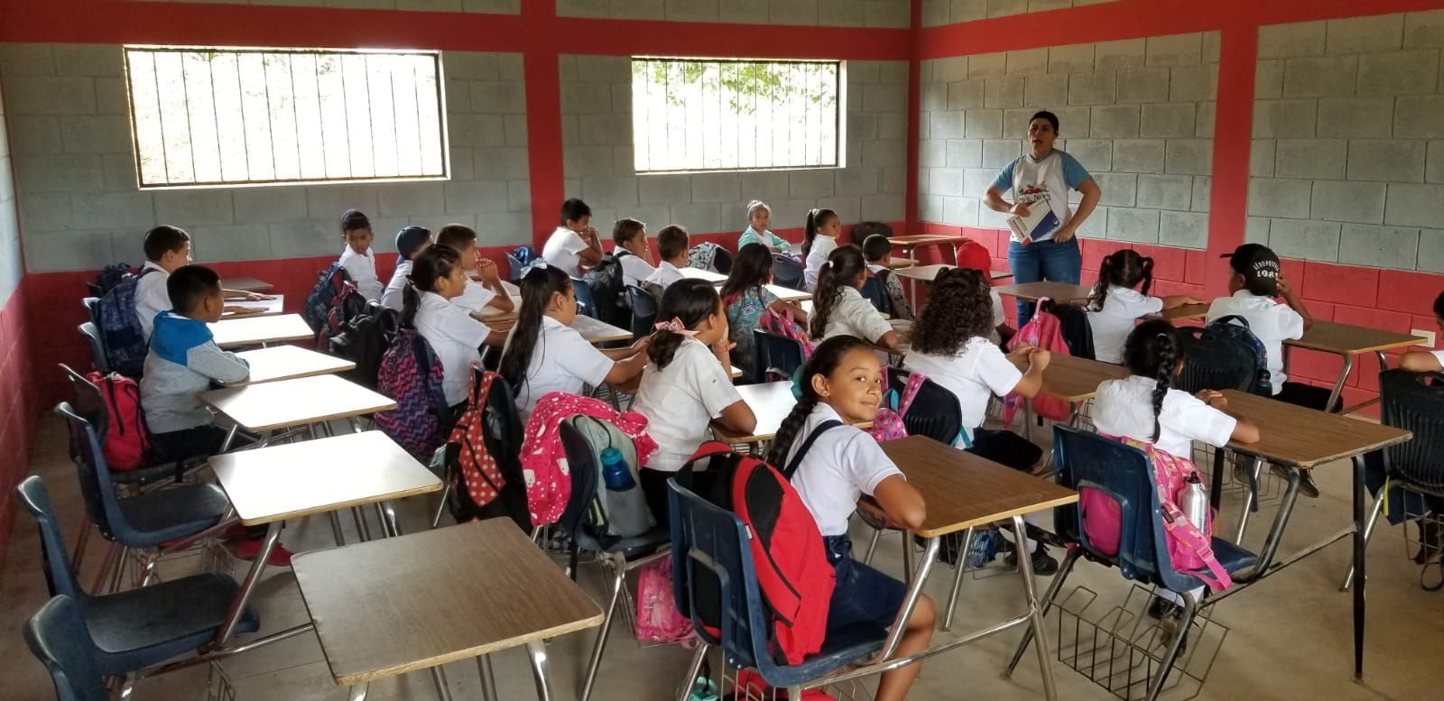 Classroom with students seated at desks listening to a teacher at the front, red walls and whiteboard visible