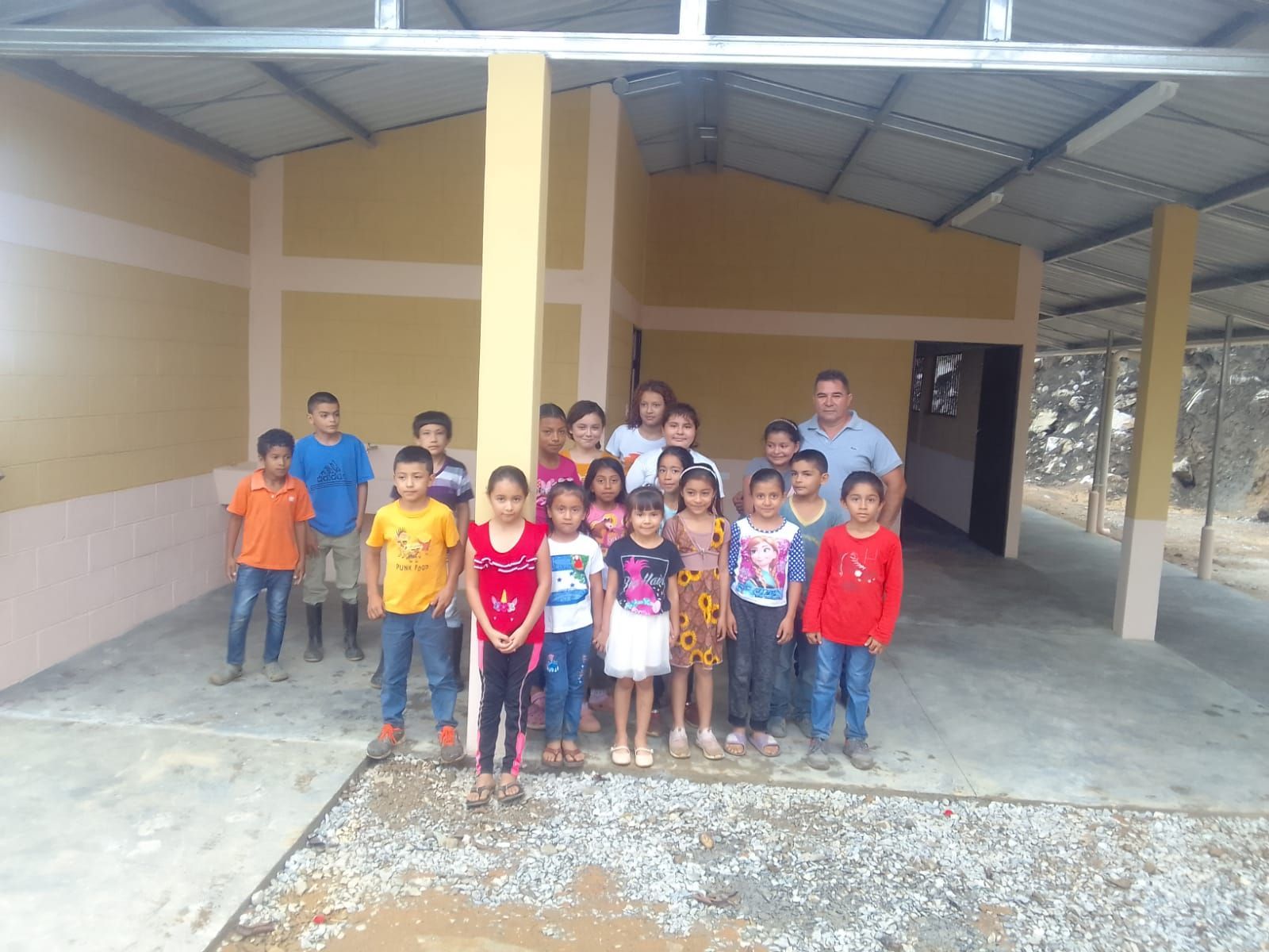 Group of children and a few adults standing under a covered outdoor building, facing the camera.